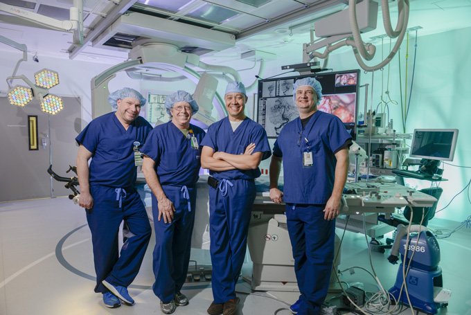 Four neurosurgeons in blue scrubs pose in an operating room