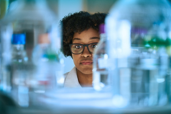 A Black woman in glasses and a lab coat is framed by glass bottles in a research lab